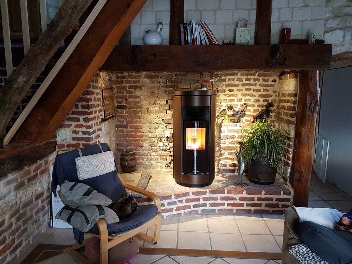 Poêle à granulés cylindrique bronze installé dans un salon rustique en briques par Hapînov